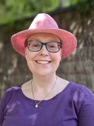 A smiling woman stands outside against a brush fence. She is wearing glasses, a pink hat and a purple top. 